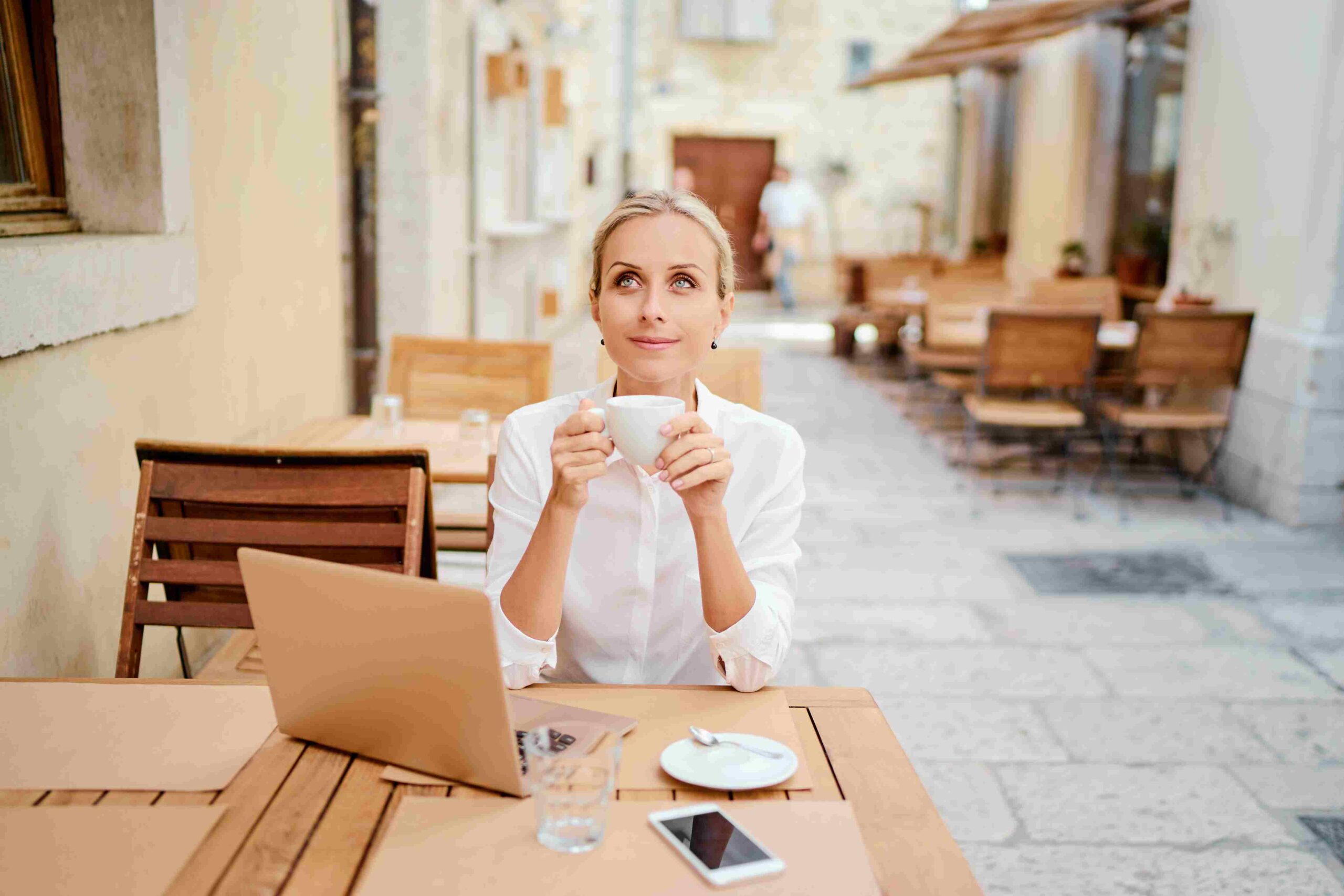 Žena sjedi u kafiću s laptopom i šoljicom kafe dok razmišlja o poslu i karijeri.